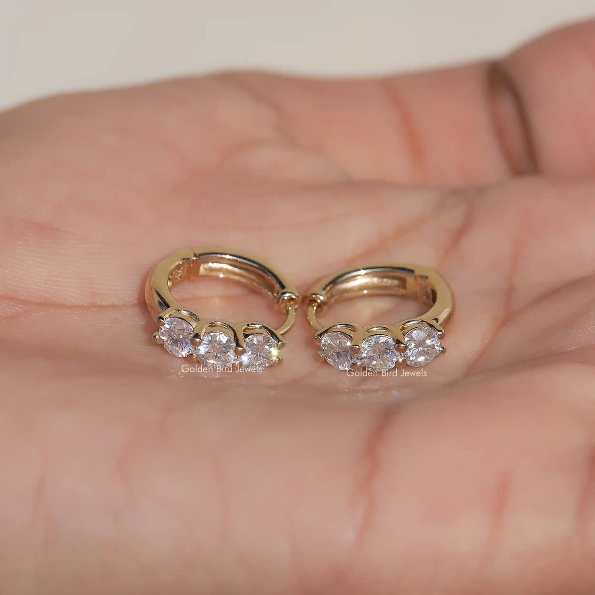 Gold-tone huggie hoop earrings with three round-cut stones, pair shown on open palm