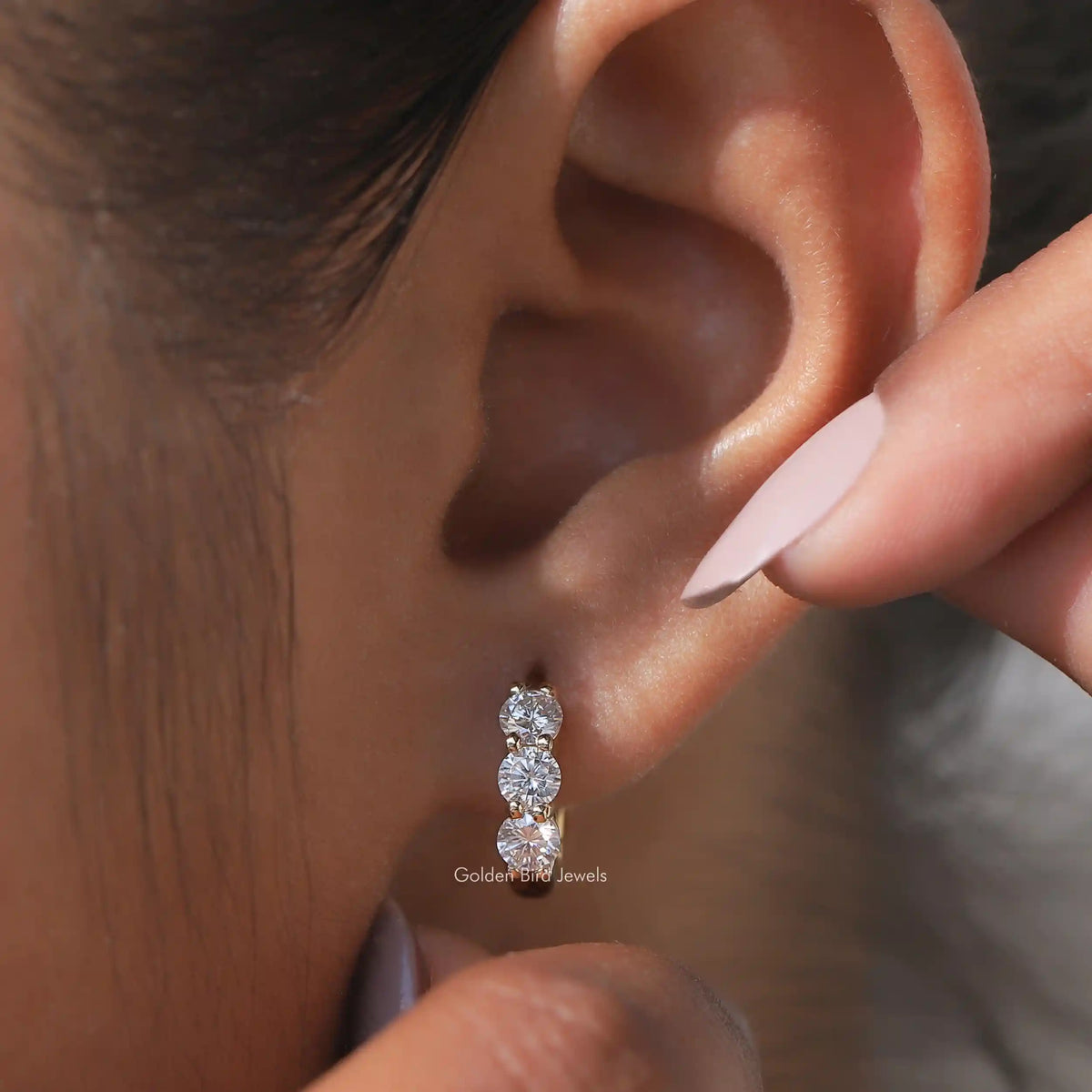 Silver-tone huggie hoop earrings with three round-cut stones, shown on ear with finger