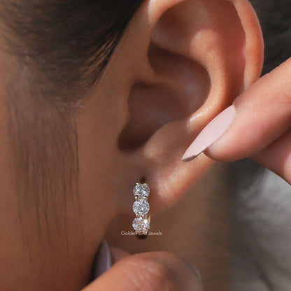 Silver-tone huggie hoop earrings with three round-cut stones, shown on ear with finger