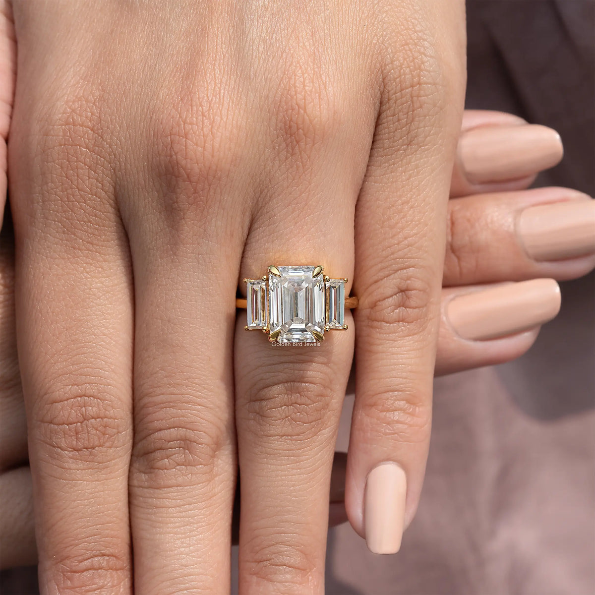 Three-stone emerald-cut moissanite ring close-up on hand, gold-tone prong setting.