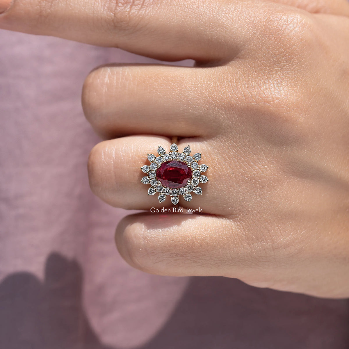 Oval red gemstone halo engagement ring with round sparkle accents, shown on finger